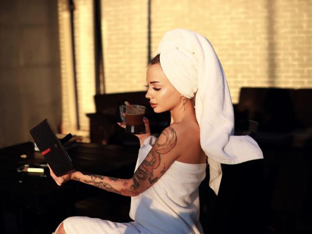 KayaBelle, con una toalla blanca en la cabeza, un brazo tatuado y el trasero cubierto con una toalla, sosteniendo un libro y una taza, sentada en una habitación con poca luz..