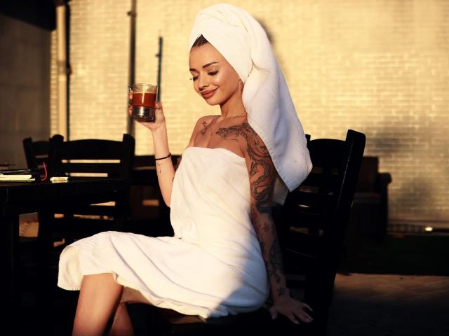 KayaBelle con una toalla alrededor de la cabeza, sosteniendo una bebida, mostrando tatuajes en el brazo y el costado, sentada en una silla, sonriendo, con el trasero desnudo, bajo la cálida luz del sol..