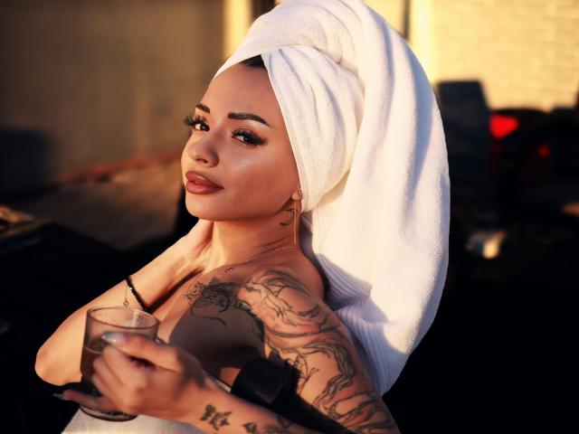 KayaBelle with a white towel on her head, holding a glass, showing tattoos on her arm, and looking at the camera with full lips and dark eyeliner.