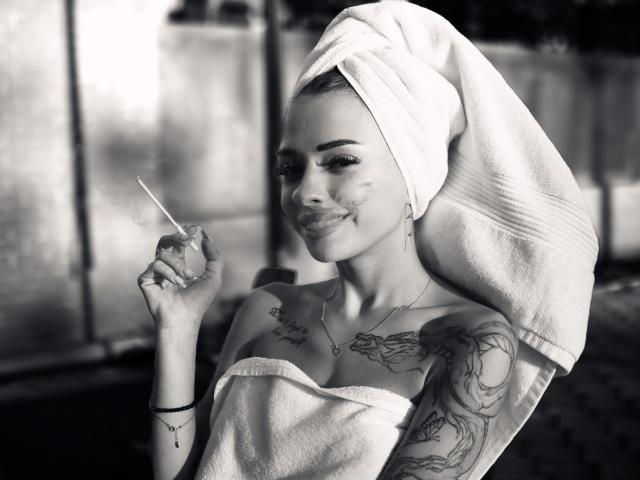 Black-and-white photo of KayaBelle with a towel on her head, holding a cotton swab, tattoos on her right arm and chest, smiling, showing facial marks, wearing a strapless towel.