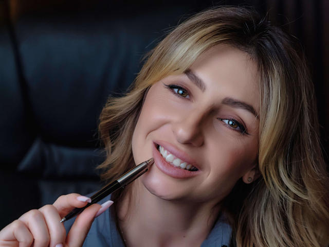 Sofia: Ragazza con i capelli biondi, sorridente, che tiene una penna sulle sue labbra rosa leggermente dischiuse, occhi azzurri, pelle chiara, indossa una camicia grigia, seduta su un divano in pelle nera..