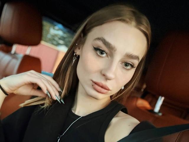 PetrinaRaven, with light skin, brown hair, and a nose ring, sits in a car, wearing black, showing off her plump lips and green eyes, with a sexy, confident look.