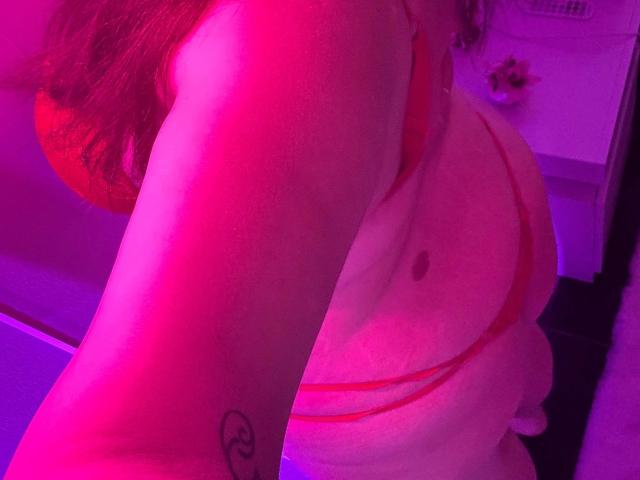 CurvyAlexaa-hots rosafarben beleuchteter, nackter Oberkörper, der kleine Brüste in einem roten, trägerlosen Top zeigt, mit einem Smiley-Tattoo auf ihrem Arm, und ihr Hintern ist teilweise sichtbar..