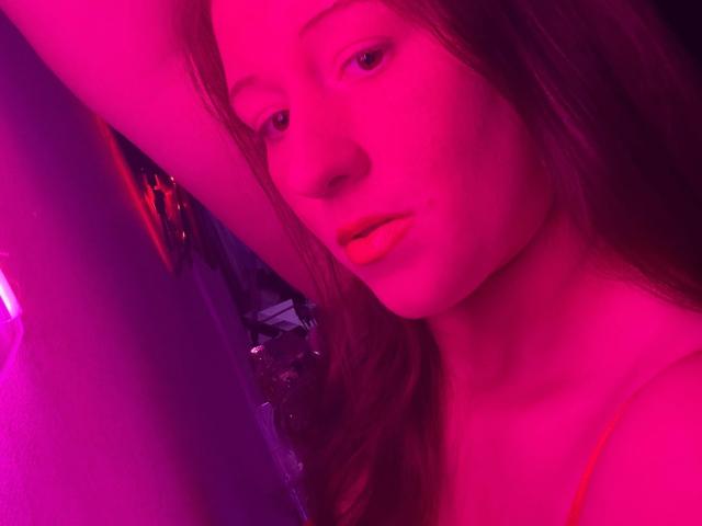 CurvyAlexaa-hot with pink-lit face, long brown hair, and orange lipstick, looking slightly bored. Her arm is raised, and she's wearing a thin-strapped top.