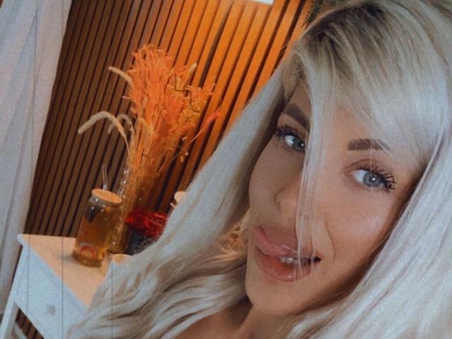 SalmaRoze a de longs cheveux blonds platine, des yeux bleus, et elle tire la langue en laissant légèrement entrouvertes ses lèvres, ce qui montre ses dents. Un verre contenant un liquide ambré est posé sur une table blanche..