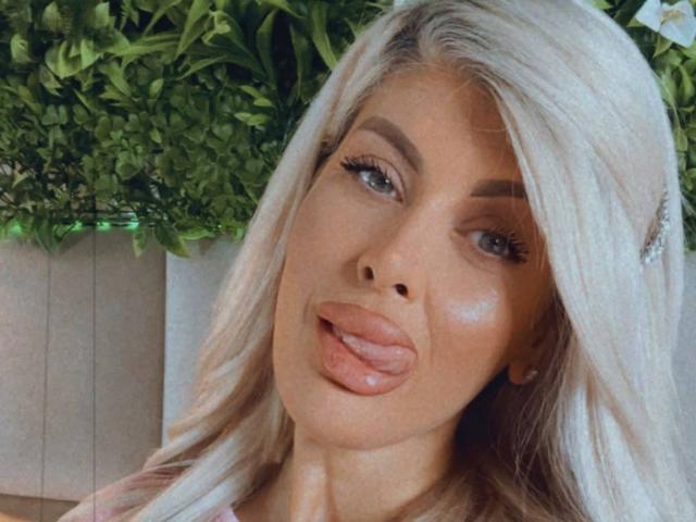 SalmaRoze, com cabelo loiro platinado, olhos azuis e uma língua rosa brilhante para fora. Ela tem lábios cheios, sobrancelhas arqueadas e um leve bronzeado. Seus cílios são longos e curvados..