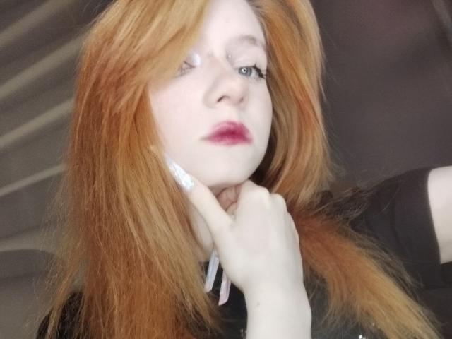 JamieLathrum has pale skin, red hair, and is touching her neck with a hand showing white nails. She has pink lipstick and a slightly wistful expression.