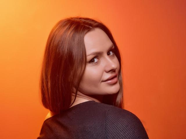 MillaVogue's brown hair, fair skin, and subtle smile are against an orange background. She's looking over her shoulder, wearing a dark, ribbed top, showing her ass.