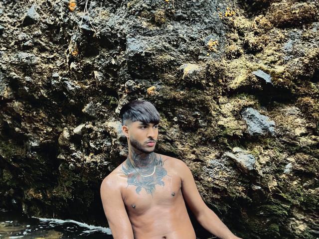 TheImperialMen, shirtless, with dark tattoos on the neck and chest, standing in front of a rocky, moss-covered wall, has a medium brown skin tone and short black hair.