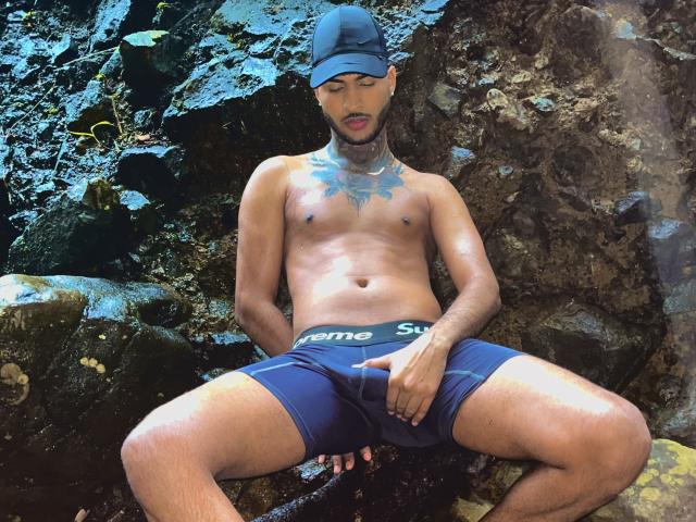 TheImperialMen, shirtless, wearing blue "reme" shorts, sits against rocks with a cap on, hand over his crotch, tattoo on chest, water droplets on skin, looking down.