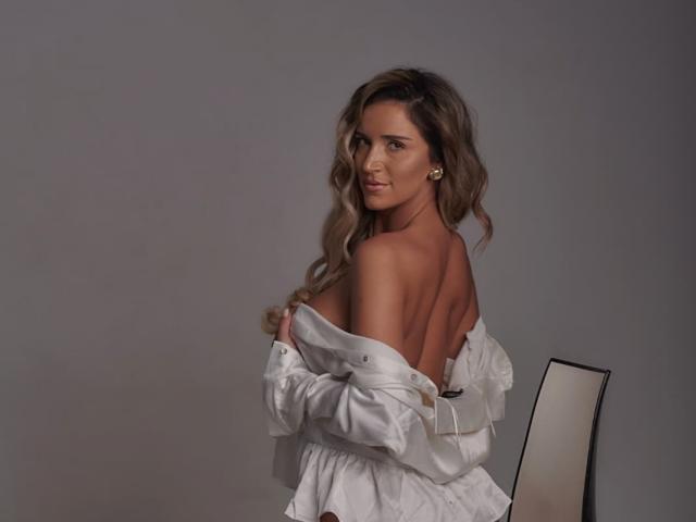 Tiffany Addams mit welligem, braunem Haar, weißem, schulterfreiem Hemd, blickt nach hinten, wobei ihr nackter Rücken und Hintern zu sehen sind, sitzt auf einem Stuhl, mit einem sanften Lächeln, vor einem grauen Hintergrund..