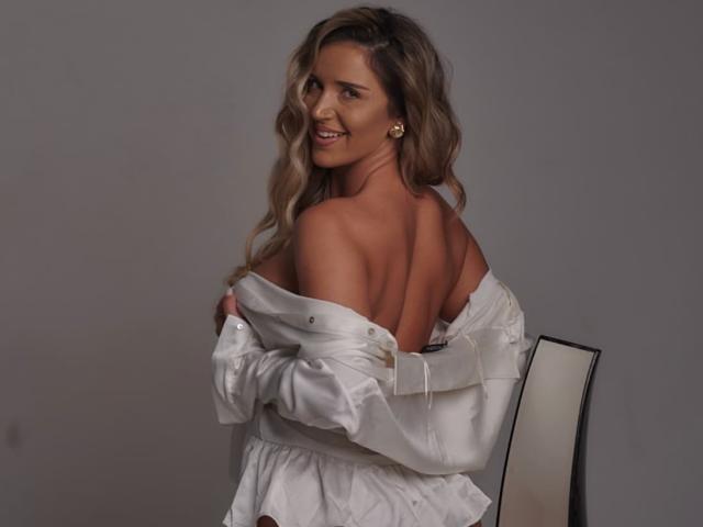 Tiffany Addams mit welligem, blondem Haar, lächelnd, trägt ein weißes Oberteil mit einem Schulterauschnitt, das ihren nackten Rücken und ihre Brüste freilegt, sitzt auf einem Stuhl und blickt über ihre Schulter..