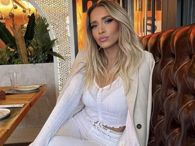Tiffany Addams mit langen blonden Haaren, pinkem Lippenstift, einem weißen Oberteil und einer weißen Hose, sitzt in einer braunen Lederecke, trägt einen beigen Mantel und blickt in die Kamera..