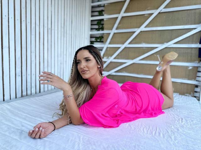 TiffanyAddams, liggend op een wit bed, gekleed in een felroze jurk, haar billen omhoog, benen gebogen, naakte hakken, lang blond haar, lachend, rechterhand omhoog, linkerhand op het bed..