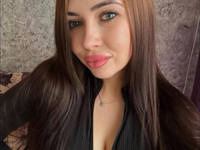 RoseKarolineI a de longs cheveux bruns, des yeux verts, et porte du rouge à lèvres rose avec un haut noir qui dévoile légèrement sa poitrine. Elle sourit doucement à l'appareil photo..