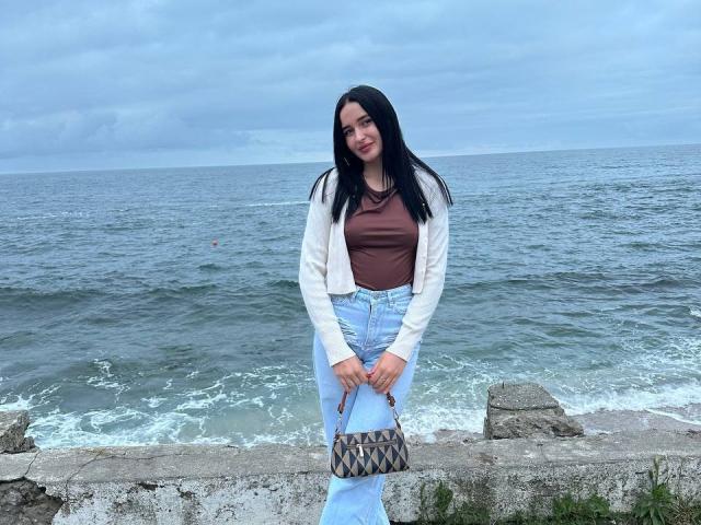NanaCherish, con lunghi capelli neri, una maglietta marrone, una giacca bianca, jeans blu, che tiene una borsa con un motivo a diamante, in piedi vicino all'oceano, che guarda leggermente verso il basso con un sorriso appena accennato..
