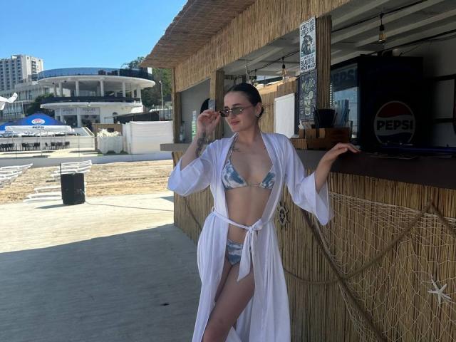 NanaCherish with glasses, white robe, blue-gray bikini, holding a drink, standing by a bamboo bar, clear sky, sunlight, short brown hair, small tits, slender, tattoo on chest.