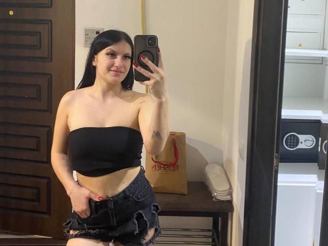 Karolina, con un top nero senza spalline che mette in mostra il seno, e pantaloncini di jeans neri strappati, sta scattando un selfie allo specchio con il suo telefono, sorridendo, con lunghi capelli neri e un tatuaggio sul braccio..