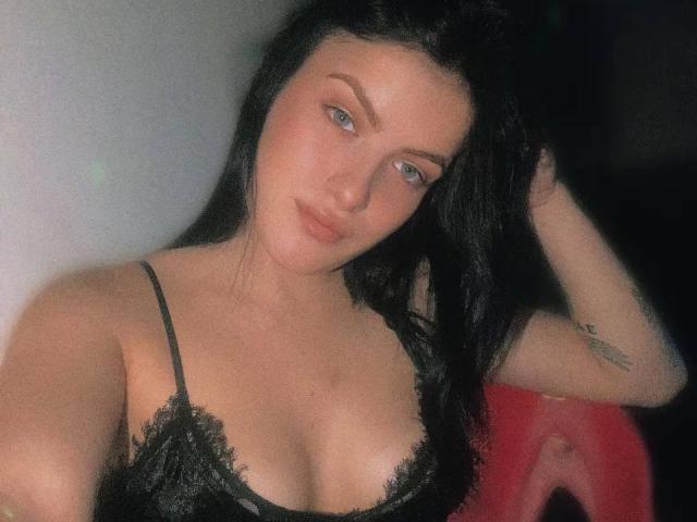 KarolinaAlegras with black lace bra, blue eyes, and long black hair, hand in hair, tattoo on right arm, medium tits, slight smile, soft lighting, red chair.