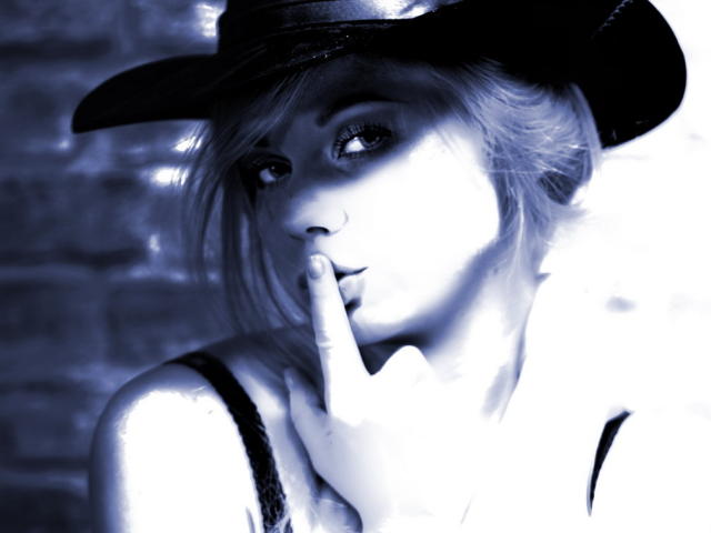 TinaRey69 mit einem glänzenden schwarzen Cowboyhut, einem blau schimmernden Gesicht und einem Finger am Mund, trägt eine schwarze Spitzenbluse, hat kurze blonde Haare und einen verführerischen, nachdenklichen Blick..