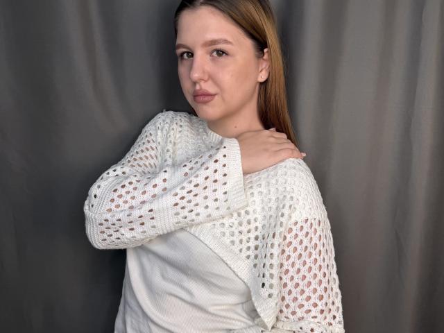Jacquetta Henneberg, con lunghi capelli castani, indossa un maglione bianco trasparente e una camicia bianca, ed è in piedi di fronte a una tenda grigia, guardando la fotocamera con un leggero sorriso..