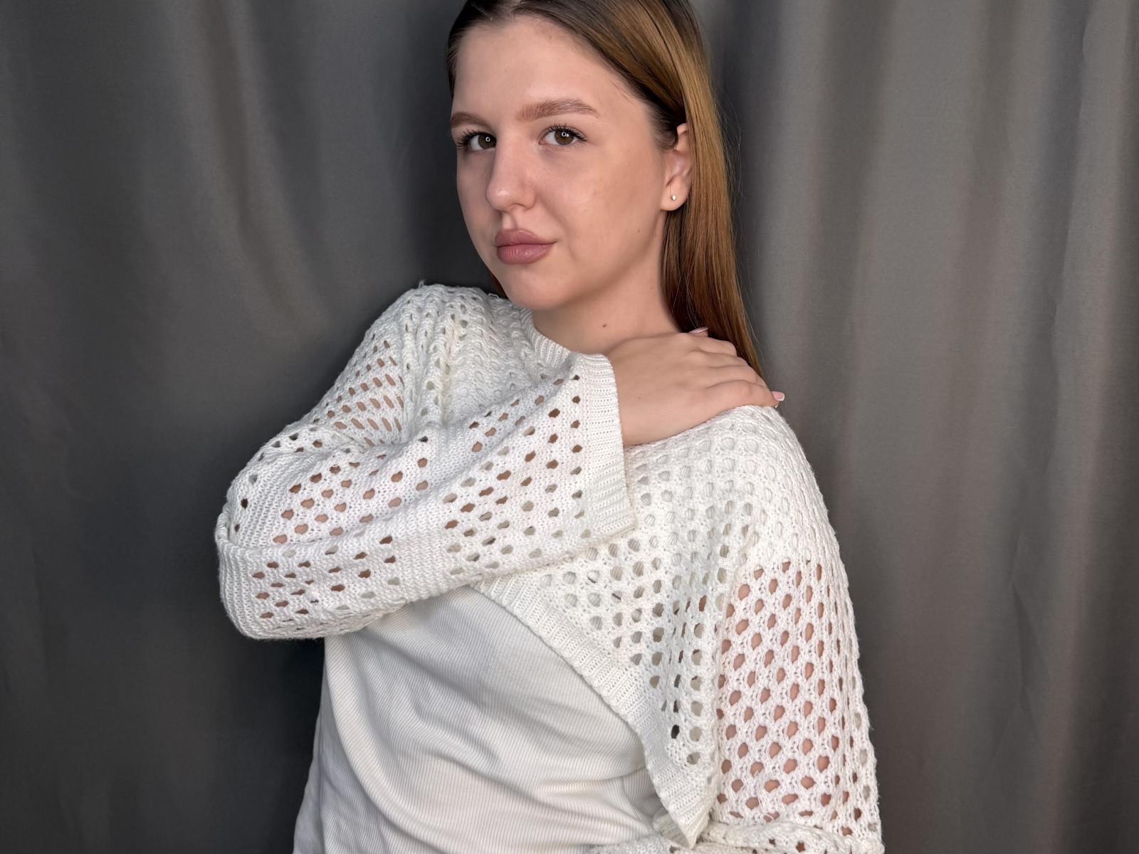 Jacquetta Henneberg, con lunghi capelli castani, indossa un maglione bianco trasparente e una camicia bianca, ed è in piedi di fronte a una tenda grigia, guardando la fotocamera con un leggero sorriso..