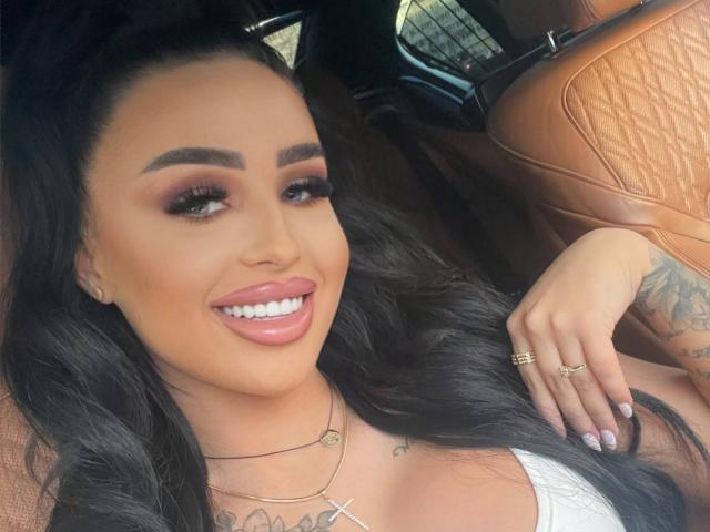 CelesteNyx with long dark hair, brown eyes, and full pink lips, smiling in a car with tan leather seats, wearing a white top, showing tattoos, and gold rings.
