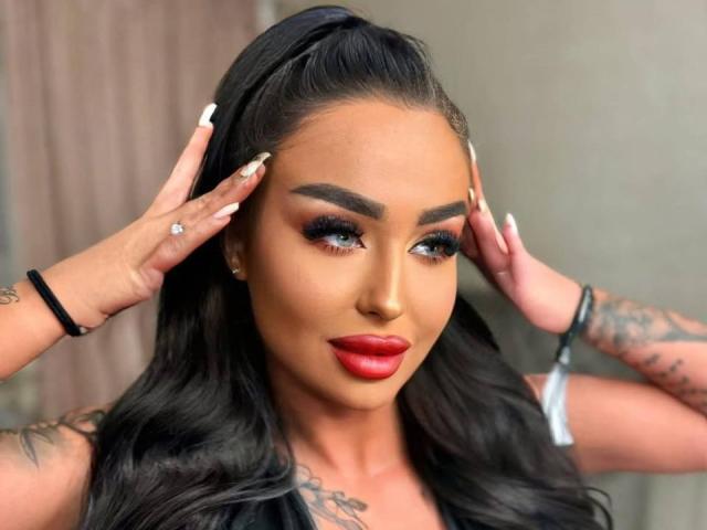 CelesteNyx con cabello oscuro en una coleta alta, lápiz labial rojo, delineador de ojos intenso, tatuajes en los brazos, manos tocando la cabeza, uñas largas, anillo en la mano derecha, pulsera en la muñeca izquierda..