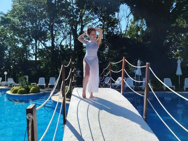 LovelyxEmilly standing on a concrete bridge over a blue pool, wearing a sheer white dress, arms raised, revealing her pussy through the fabric, sunlight casting shadows.