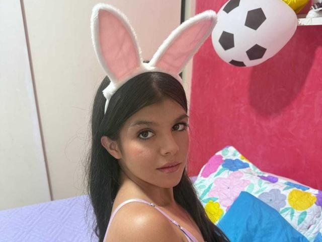 RilleyFoyx with long black hair, wearing white bunny ears, looking back over her shoulder, in a pink room, with a soccer ball and colorful pillow. Boobs not visible.
