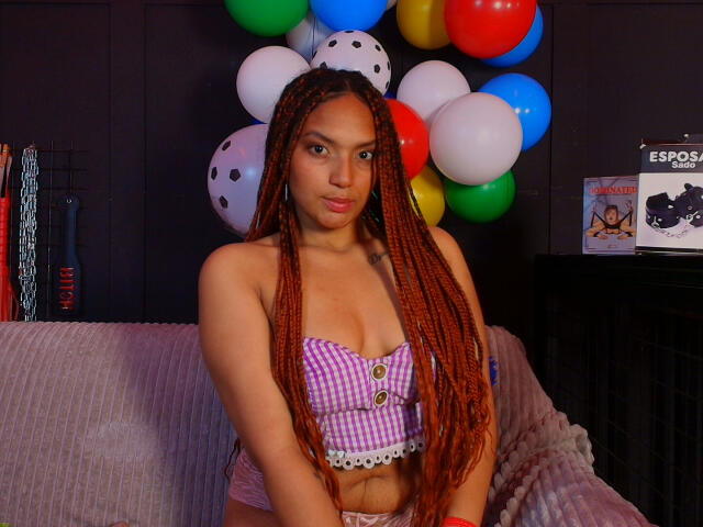 BrianaPratt with long red braids, wearing a purple checkered top that shows her small tits, standing in front of colorful balloons, looking directly at the camera.