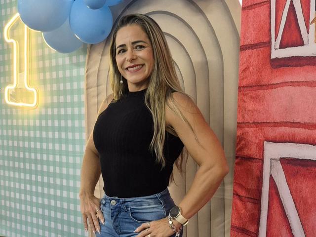 CarollineBer with long blonde hair, black sleeveless top, blue jeans, smiling, hands on hips, toned arms, visible breasts, wristwatch, standing in front of colorful background.