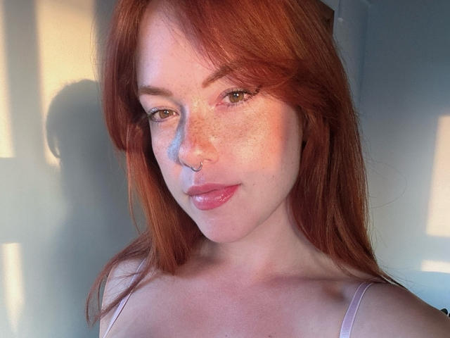 SophyeEsra tiene el pelo rojo, pecas y un piercing en la nariz. Está mirando a la cámara con los labios rosados, lleva una blusa rosa y tiene una sombra a su derecha..