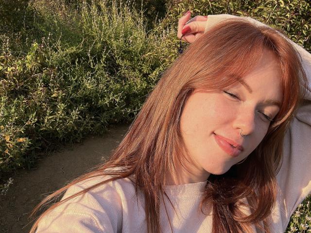 SophyeEsra, com cabelo ruivo, olhos fechados, piercing no nariz e um sorriso discreto, vestindo uma camisa branca, com o braço levantado, luz do sol no rosto, em pé em uma área gramada..