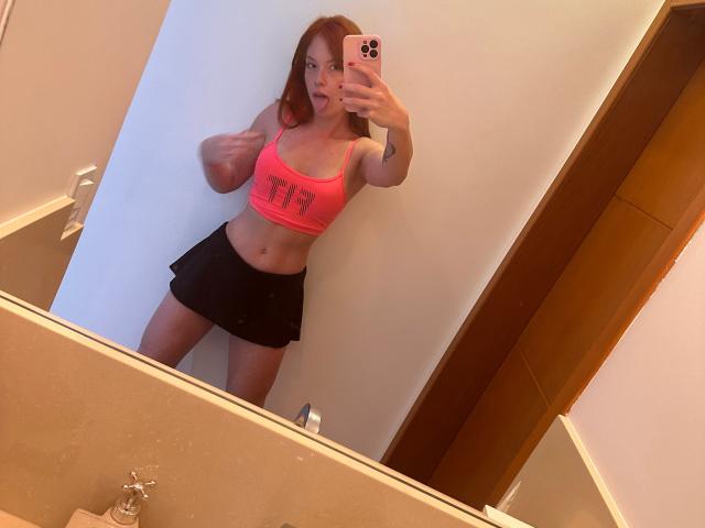 SophyeEsra met rood haar, een roze sportbeha, die haar spieren laat zien, haar tong uitsteekt, een zwarte mini-rok draagt en een spiegel-selfie maakt in een badkamer..