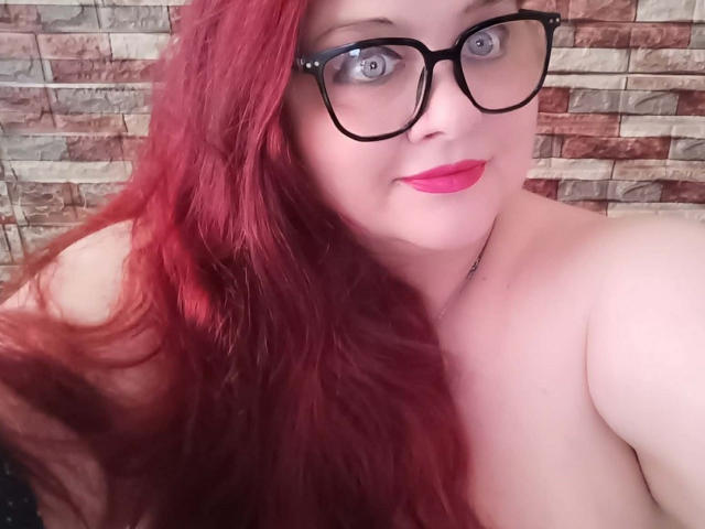 MolligeDani mit roten Haaren, schwarzen Brillen und pinkem Lippenstift, lächelnd, mit freiliegenden Schultern, blauen Augen und heller Haut, hält ihr Haar auf der linken Seite..