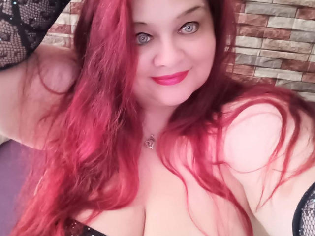 MolligeDani, con cabello rojo, ojos azules y lápiz labial rosa, sonriendo a la cámara, vestida con encaje negro, mostrando sus grandes senos y un collar con forma de corazón..