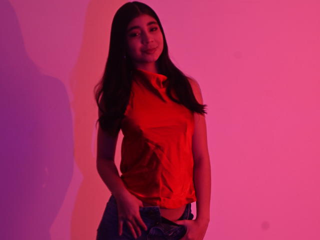 LyzMoon, avec ses longs cheveux noirs, portant un haut rouge sans manches et un jean bleu, se tient devant un éclairage rose-violet, souriant doucement, ses mains reposant sur son jean..