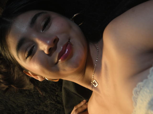 El rostro de LyzMoon, con labios rosados brillantes, una nariz pequeña y cejas oscuras, lleva un collar con un colgante en forma de corazón, y está acostada con encaje blanco visible en su pecho..