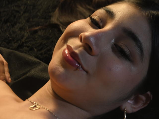 Le visage de LyzMoon, les yeux fermés, affiche un sourire avec des lèvres roses brillantes, un maquillage discret et un petit piercing au nez. Elle porte un collier en or et des boucles d'oreilles créoles, et est allongée..