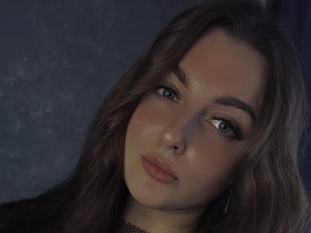 KellyJeanie's face, showing full lips, blue eyes, and long brown hair, with a slight smile and subtle makeup, in a soft, dimly-lit portrait.