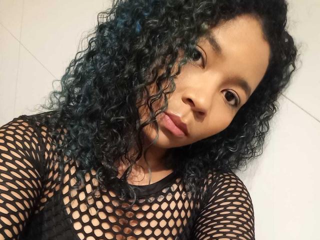 XinemaBonet mit lockigem, blau-grünem Haar, trägt eine schwarze Netzoberteil, zeigt kleine Brüste und hat braune Augen, blickt mit neutralem Ausdruck in die Kamera..