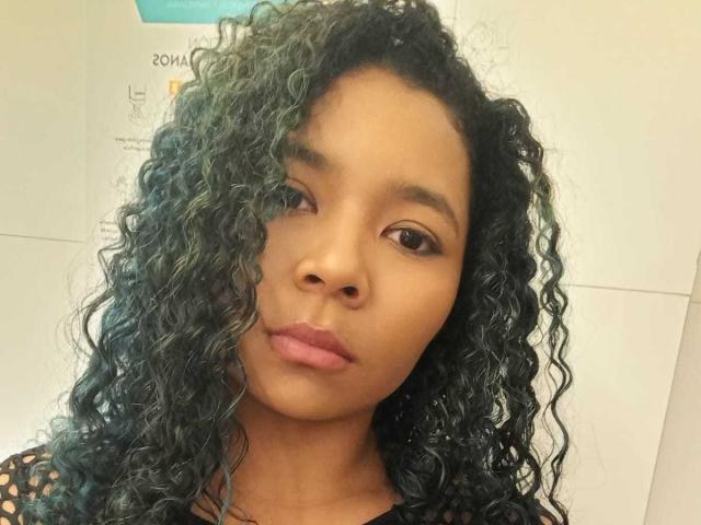 XinemaBonet tiene la piel de color marrón oscuro, cabello rizado de color verde azulado y labios rosados, y lleva una blusa negra de encaje, con una expresión seria y una ligera sonrisa..