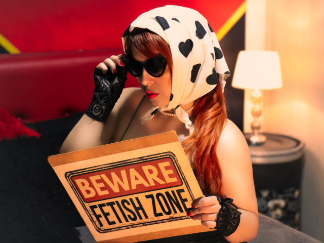 Lilibel with red hair, black sunglasses, cow print hood, black lace gloves, holding "Beware Fresh Zone" sign, sitting on bed, wearing red lipstick, sexy and confident.
