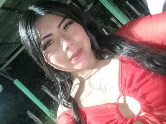 CataMoreno, con i suoi lunghi capelli neri, indossa un abito rosso con un taglio che lascia intravedere parte del corpo, ha un viso carino e un leggero sorriso, e i suoi seni sono parzialmente visibili attraverso l'abito..