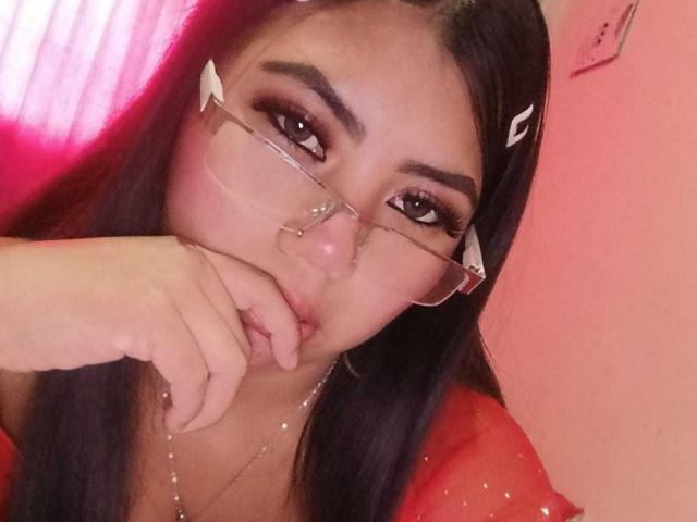 CataMoreno with long black hair, wearing glasses and a red top, has her finger near her pouty lips, looking seductive with a necklace and pinkish lighting.