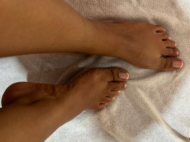 NatalyBlonder's bare feet with brown skin and pink toenails, toes slightly curled, lying on a white textured surface. Her toes are neatly trimmed and her nails are polished.