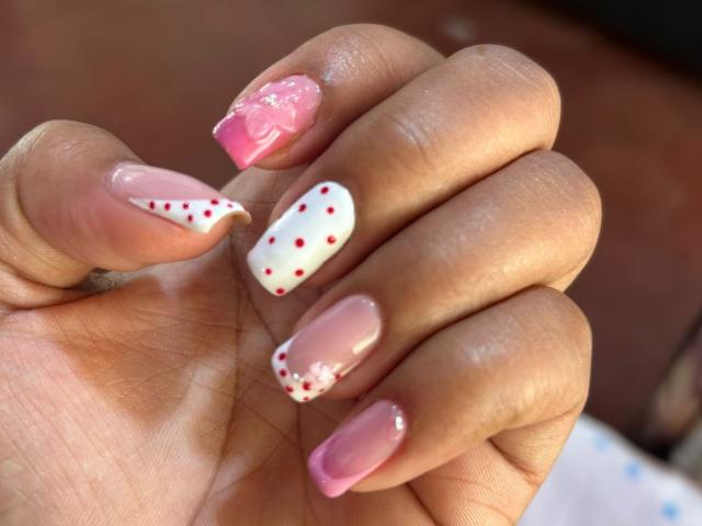 La main de NatalyBlonder, avec des ongles roses brillants, des ongles blancs avec des pois rouges, et des pointes roses, présente de jolis motifs floraux blancs et roses, mettant en valeur sa manucure élégante..