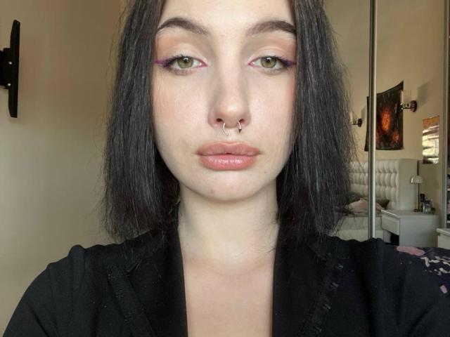 LavonaLenzo ha la pelle chiara, occhi verdi con eyeliner rosa, un piercing al naso e capelli neri lunghi fino alle spalle. Indossa una maglietta nera e guarda direttamente verso la fotocamera..