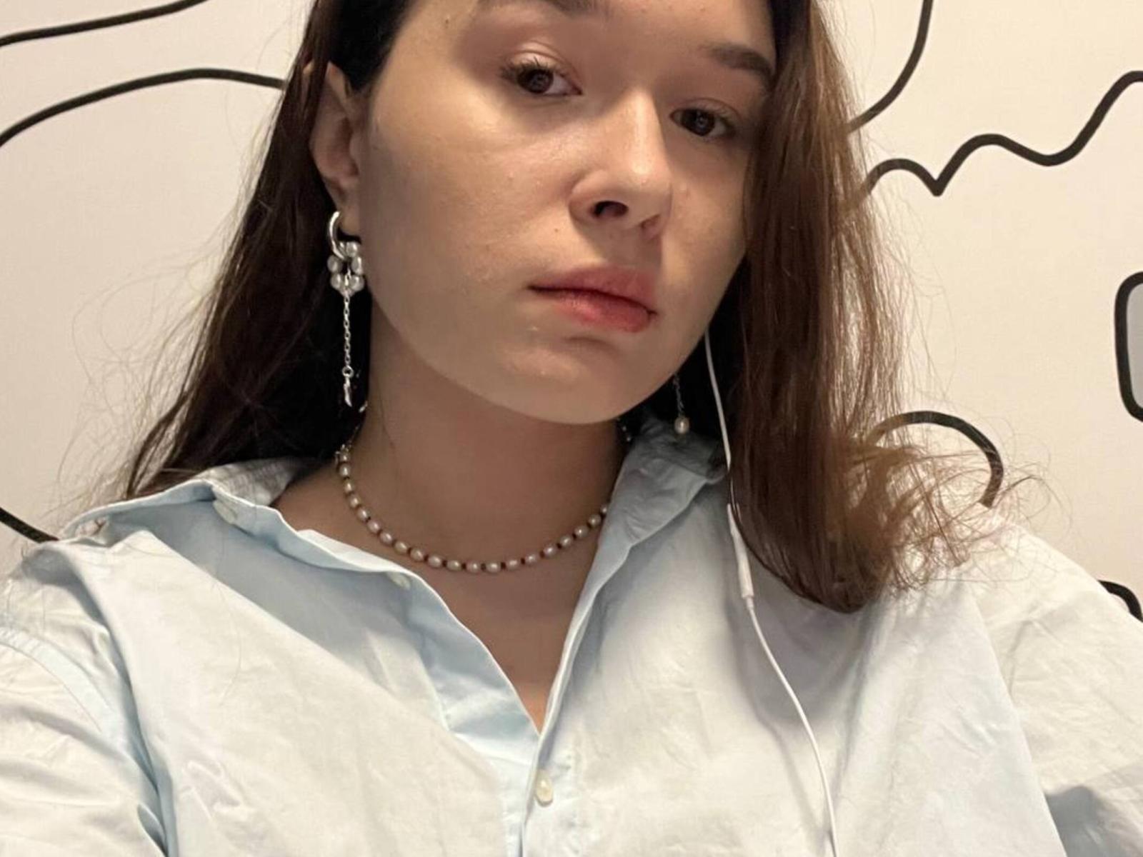 Jannet Rundahl, mit langen braunen Haaren, trägt ein hellblaues Hemd mit Knopfleiste, eine Perlenkette und Ohrringe mit Blumen. Sie hat einen ernsten Gesichtsausdruck und ein Headset im rechten Ohr..